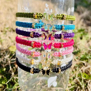 May include: A collection of colorful macrame bracelets with butterfly charms. The bracelets are in various colors, including green, blue, purple, pink, white, and black. Each bracelet features a gold-colored clasp and butterfly pendant.