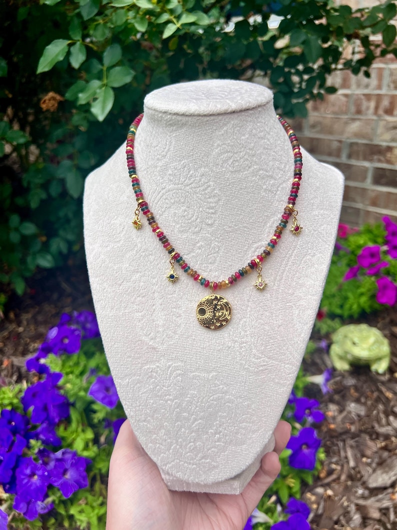 May include: A beaded necklace featuring small, multi-colored gemstone rondelles in shades of red, green, and yellow. The necklace includes four gold-toned charms: three small star-shaped pendants and one larger circular pendant depicting a sun and moon.