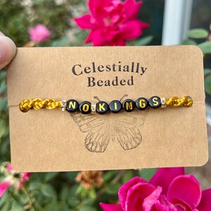 May include: A gold-colored beaded bracelet with black letter beads spelling "NOKINGS" is displayed on a brown card. The card is labeled "Celestially Beaded" and features a butterfly graphic. The background includes pink flowers.