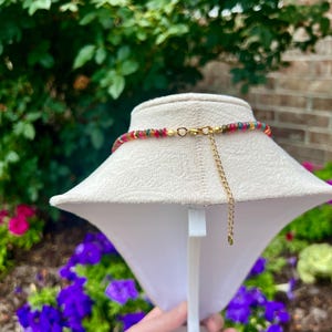 May include: A necklace featuring a strand of small, multi-colored beads in shades of red, yellow, green, and blue. The necklace includes a gold-toned metal clasp and a short gold-toned extender chain hanging from the back of the display stand.