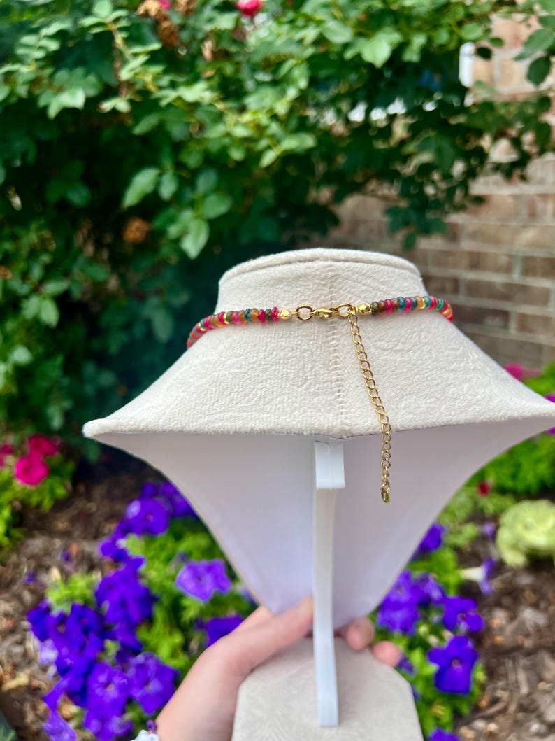 May include: A necklace featuring a strand of small, multi-colored beads in shades of red, yellow, green, and blue. The necklace includes a gold-toned metal clasp and a short gold-toned extender chain hanging from the back of the display stand.