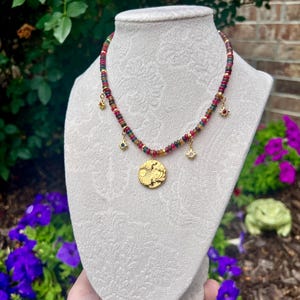 May include: A necklace featuring a strand of small, multicolored beads in shades of red, green, and yellow. The necklace includes four gold-toned charms: three small star-shaped pendants and one larger, circular pendant embossed with a sun and moon design.