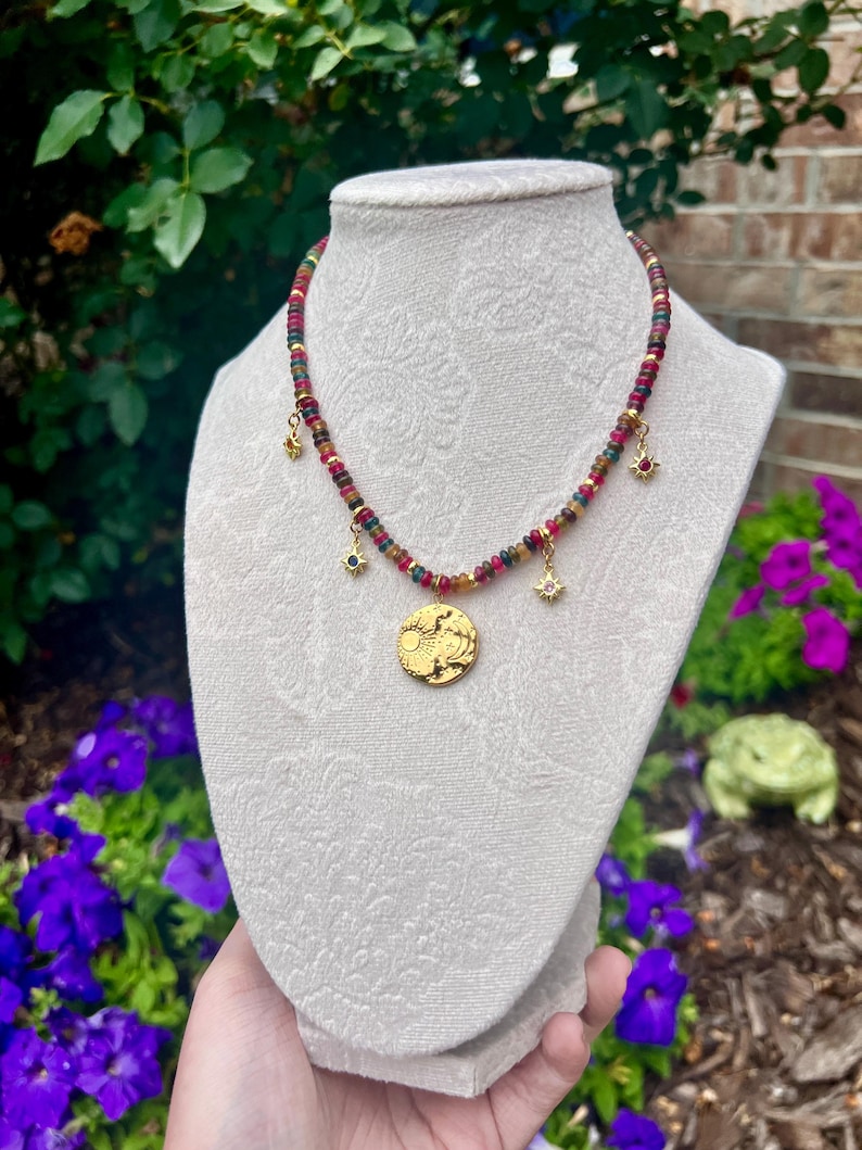 May include: A necklace featuring a strand of small, multicolored beads in shades of red, green, and yellow. The necklace includes four gold-toned charms: three small star-shaped pendants and one larger, circular pendant embossed with a sun and moon design.