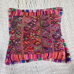 Puede incluir: Un cojín cuadrado y vibrante con un complejo patrón geométrico. El cojín presenta un diseño de patchwork con una variedad de colores que incluyen rojo, rosa, morado, verde y amarillo. Los bordes están adornados con un borde multicolor.