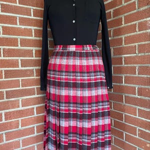May include: A long-sleeved black button-down shirt with a collar and a single chest pocket paired with a high-waisted, pleated midi skirt featuring a vibrant plaid pattern in shades of bright red, light gray, and dark charcoal gray throughout.