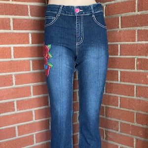 May include: Dark blue denim jeans featuring a high-waisted fit with a bright pink button closure. The front of the right leg displays a colorful embroidered graphic of a red rose with green leaves and blue accents against the dark fabric.