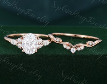 May include: A rose gold engagement ring set. The engagement ring features an oval-cut center stone surrounded by smaller stones. The matching band has a curved design with delicate stone accents. The rings are displayed on a dark green background.