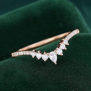 May include: A rose gold V-shaped ring featuring a central pear-cut diamond flanked by a series of smaller round-cut diamonds along the band. The band has a polished finish and follows a curved contour, showcasing a delicate and symmetrical design.