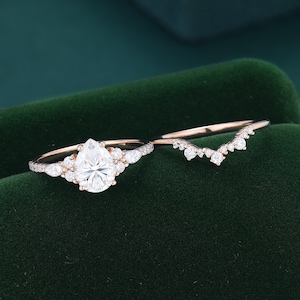 May include: A rose gold engagement ring set featuring a pear-cut center stone flanked by smaller round and marquise-cut stones, paired with a matching V-shaped chevron band adorned with a series of small round stones along the thin metal shank.