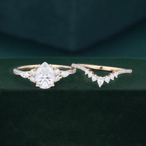 May include: A gold engagement ring set featuring a pear-cut center stone flanked by smaller round and pear-shaped diamonds on a thin band, paired with a matching curved wedding band adorned with a row of round diamonds and a central pear-cut stone.