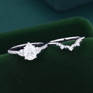 May include: A silver-toned ring set featuring a pear-shaped center stone flanked by small round stones on a pavé band, paired with a matching chevron-shaped band adorned with a series of small round stones along the top edge of the metal.