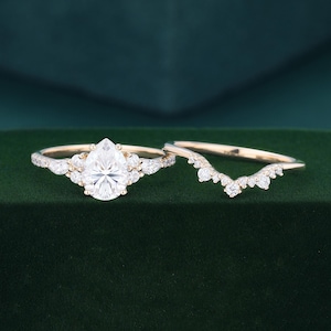 May include: A gold engagement ring set featuring a central pear-cut stone flanked by smaller round and marquise-cut stones, paired with a matching curved wedding band adorned with a series of small round stones along the top edge.
