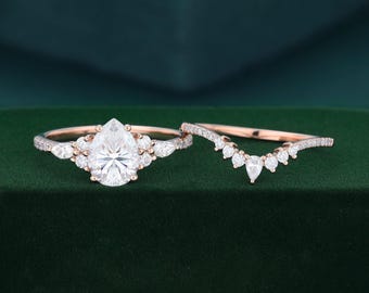 Puede incluir: Un conjunto de anillos de compromiso en oro rosa con una piedra central en forma de pera flanqueada por piedras más pequeñas redondas y en forma de pera, junto a una alianza curva a juego adornada con una hilera de pequeñas piedras.