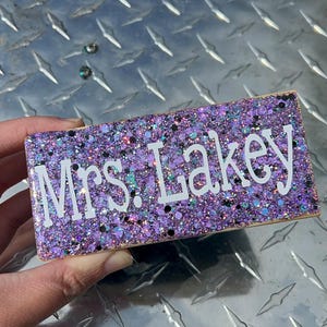 May include: A purple glitter block with the text "Mrs. Lakey" in white lettering.