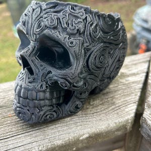 May include: A black decorative skull with intricate, swirling patterns. The skull is 3D-printed and has a hollow interior. The design includes floral and geometric elements, creating a gothic or fantasy aesthetic. The skull is resting on a wooden surface.