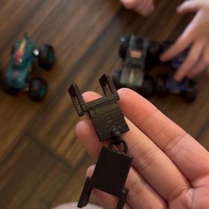 May include: A black 3D-printed toy chair held in a hand, with other toy trucks in the background. The chair has a rectangular seat and four legs. The background shows a wooden floor.