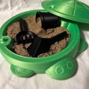 May include: A green turtle-shaped container filled with sand, featuring a lid and black accessories. The container has a round shape with a turtle shell design on the lid. Includes a small rake, scoop, and bucket.