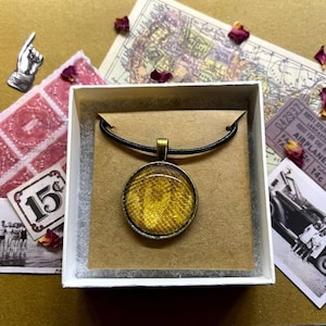 May include: A gold pendant necklace with a round, textured, yellow glass charm. The necklace is displayed in a white box with a brown paper insert.