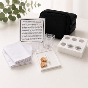 May include: A communion set featuring a black carrying case, a white tray with six circular indentations, two clear acrylic cups, a small white plate with two pieces of bread, a white cloth, and a card printed with The Blessing of the Bread prayer.