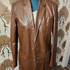 May include: A brown leather blazer with a two-button closure and two flap pockets. The blazer has a classic style and is made of high-quality leather.