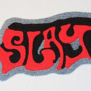 May include: Embroidered patch with the word "SLAY" in red and black, stitched onto a denim fabric backing. The patch has a wavy, irregular shape, and the text is in a bold, stylized font.