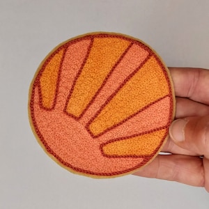 May include: A round, embroidered patch with a stylized sun design. The sun is orange and red with a textured, stitched surface.
