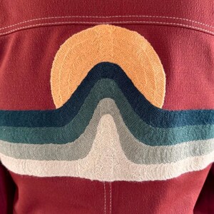 May include: Maroon jacket with an embroidered design of a sun setting over layered, wavy lines in shades of orange, green, blue, and white. The stitching is visible, adding texture to the design. The jacket has white stitching along the seams.