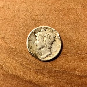 May include: A silver Mercury dime from 1943, featuring a profile of a woman with a winged Phrygian cap. The coin is slightly worn and has a visible date. The coin is on a wooden surface.