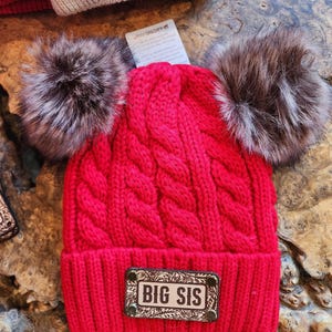 May include: A bright red knit beanie with a cable knit design, featuring two large faux fur pom-poms. The hat has a rectangular patch that reads "BIG SIS". A second beanie with "MINI" is partially visible.