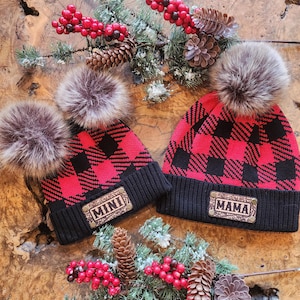 May include: Two matching red and black plaid knit hats with faux fur pom-poms. One hat has a patch that says "MINI" and the other says "MAMA". The hats are displayed on a wood surface with pinecones and berries.