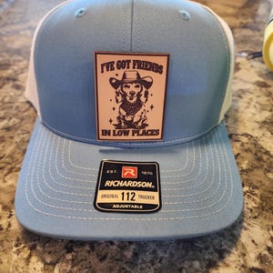 May include: A light blue trucker hat with a white mesh back. The hat features a patch with a dachshund wearing a cowboy hat and the text "I'VE GOT FRIENDS IN LOW PLACES." The hat also has a Richardson 112 Trucker tag.