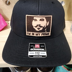 May include: Black Richardson 112 trucker hat with a square patch featuring a portrait and the phrase "I'M IN MY PRIME." The hat has a mesh back and an adjustable strap. The Richardson label is visible.