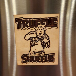 May include: Wooden square with the words "TRUFFLE SHUFFLE" in a bold font. The image features a cartoon illustration of a person with their arms raised, mouth open, and eyes wide. The design is etched into the wood.