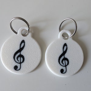 May include: Two white, circular keychains with a black treble clef design. Each keychain has a silver metal ring attached at the top. The keychains are on a white surface.