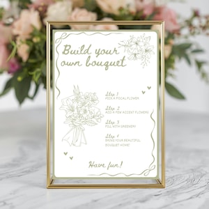 May include: A framed sign with the text "Build your own bouquet" and instructions for creating a floral arrangement. The sign is in a gold frame, with a floral illustration and the words "Have fun!"