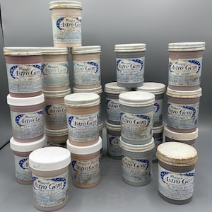 May include: A collection of Mayco Astro Gem jars in various colors, including amethyst, soft coral, gray granite, and meteor brown. The jars have white lids and labels with the Astro Gem logo and color names. The jars are stacked on a gray surface.