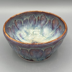 May include: A handcrafted ceramic bowl with a distinctive glaze. The bowl showcases a mix of blue, purple, and brown tones, with a shiny finish. The rim has a golden-brown accent. This decorative bowl is suitable for serving or display.