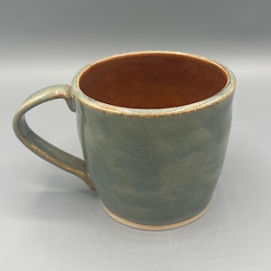May include: A handmade ceramic mug with a rounded body and a handle. The exterior is a mottled blue-green, while the interior has a warm brown glaze. The mug has a capacity of approximately 12 ounces.