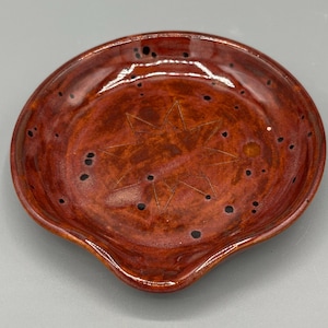 May include: A round, glossy ceramic spoon rest with a deep reddish-brown glaze and dark brown speckles throughout. The center of the dish features a simple, etched eight-pointed star design. The edges are slightly raised with a small indentation.