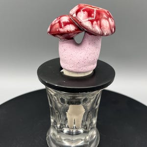 May include: A whimsical ceramic mushroom sculpture with two red and white-spotted caps atop a pink stem. The sculpture is mounted on a black base, which sits on a clear glass container. The mushroom design adds a touch of nature-inspired art.