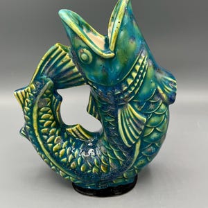 May include: A ceramic fish-shaped pitcher with an open mouth forming the spout. The piece features a glossy glaze in shades of deep teal, turquoise, and yellow, with detailed scales, fins, and a curved body resting on a circular black base.