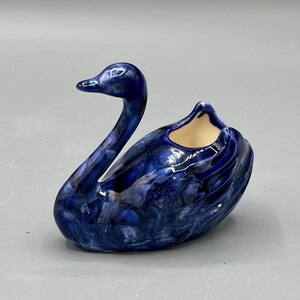 May include: A ceramic swan ornament in shades of blue with a glossy finish. The swan has a long, curved neck and a small opening on its back. The ornament is a decorative item.