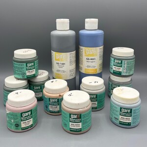 May include: A collection of GARE Simplicity non-toxic glaze bottles and jars in various colors, including blue, green, and pink. The bottles have white lids and labels with product information. The image showcases a variety of art supplies.