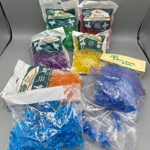 May include: Assortment of Deco Brites candle blossom beads in various colors, including green, red, purple, yellow, orange, and blue. The beads are packaged in clear plastic bags with labels from Tinker Creek Crafts.