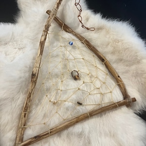 May include: A triangular dream catcher made of natural wood branches with a white string net. The dream catcher has a blue and white bead, a wooden bead, and a metal ring hanging from it.