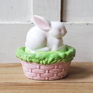 May include: A white ceramic bunny figurine with pink inner ears sits in a pink basket with green grass trim. The Easter-themed decoration is set on a wooden surface against a white paneled background.