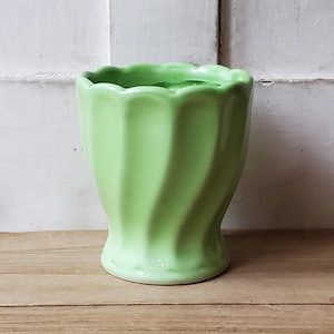 May include: A light green ceramic vase with a wavy, spiral design. The vase has a scalloped rim and sits on a wooden surface. The background is a white, weathered wooden door.