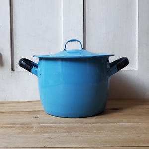 May include: A light blue enamel pot with a matching lid. The pot has two black handles and a black handle on the lid. The pot is sitting on a wooden surface.