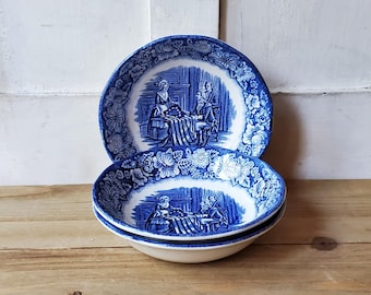 May include: Two blue and white ceramic bowls stacked on a wooden surface. The bowls feature a floral border and a central scene of people seated around a table. The bowls are a vintage style.