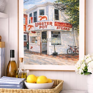 May include: A framed print depicting a white building with red signage reading "Lobster Pot Charcoal Broiled Steaks." A red bicycle is parked outside. The image is displayed on a white countertop with a woven tray holding lemons and a bowl.